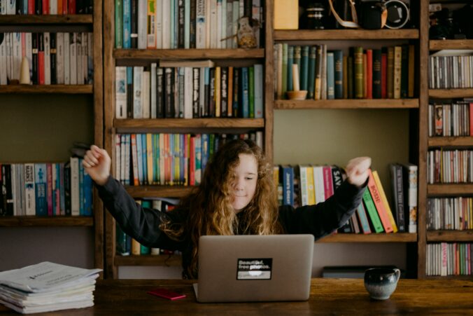 Image of a young girl with long brown hair with her hands up in the air at her sides while working on her laptop computer on a table infront of a bookcase full of books