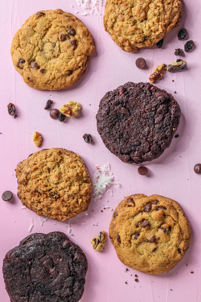 Image of 6 chocolate chip cookies, 1/3 of which are dark chocolate. The cookies are on a purple background with loose chocolate chip ingredients sprinkled around them. 