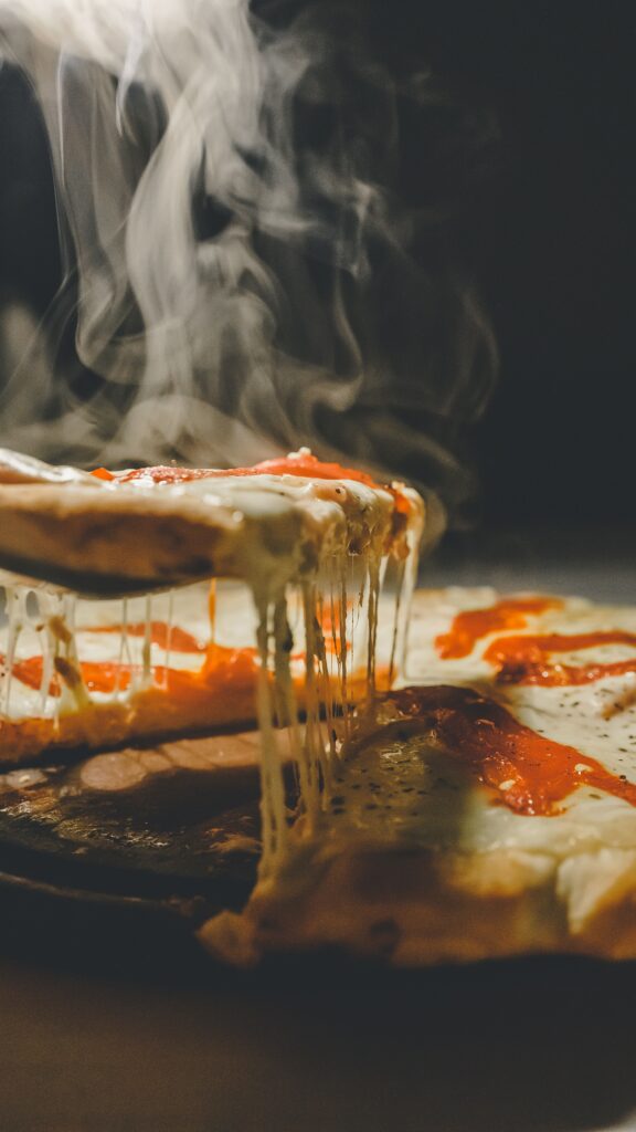 Image of a steaming hot pizza with white and red sauce. The pizza is close up on a counter and one slice is being picked up as gooey cheese melts down the sides.